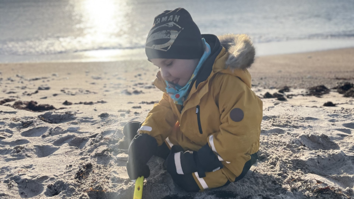 Caspian sitter p&aring; stranden, med jacka, vantar och m&ouml;ssa och gr&auml;ver